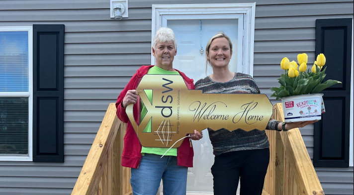 Two women standing on a wooden porch holding signs that say "Welcome Home" and a potted plant with yellow tulips.