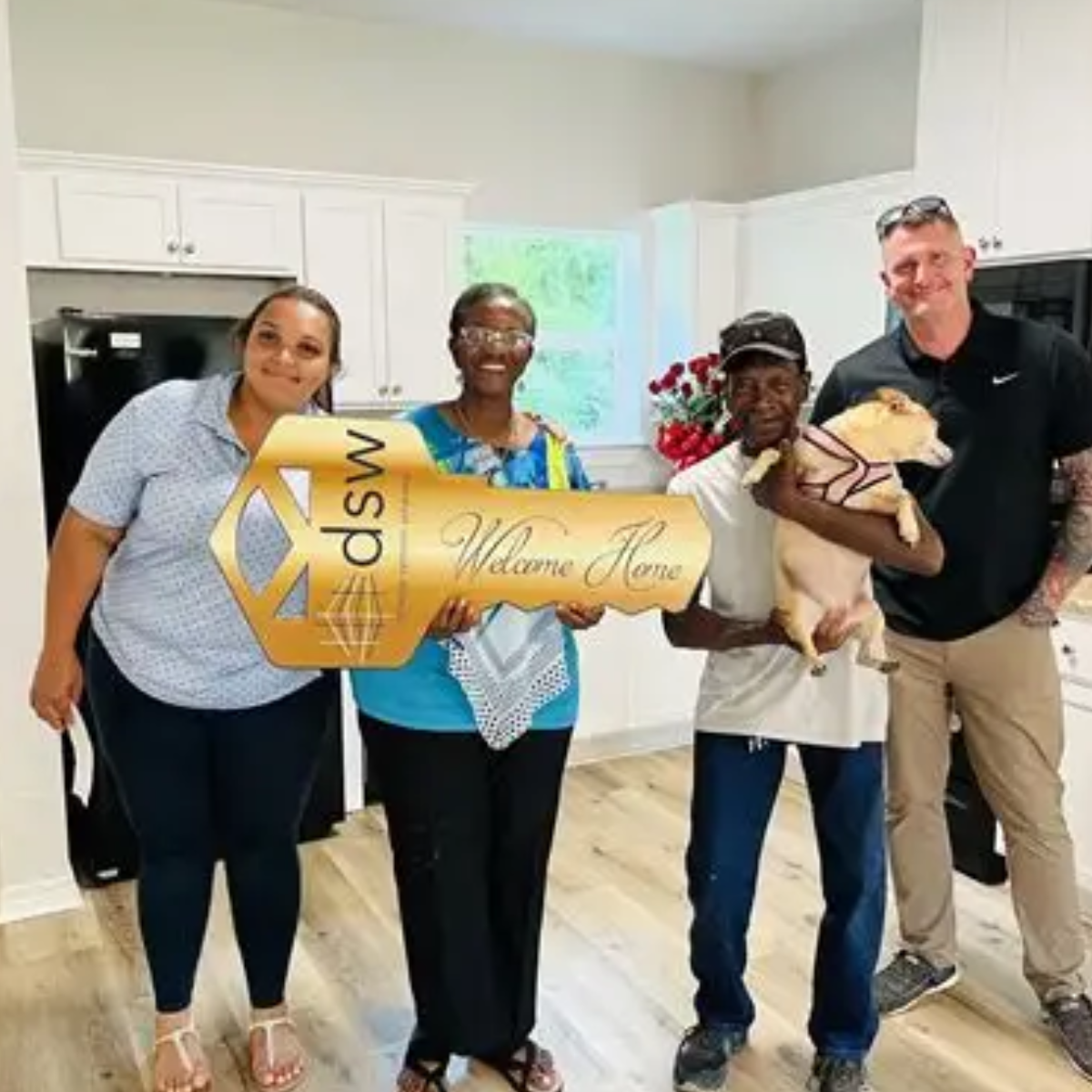 Group of four adults in a kitchen holding a large golden sign that reads 'dsm' and 'Welcome Home', with one person holding a small dog. They are smiling and celebrating.