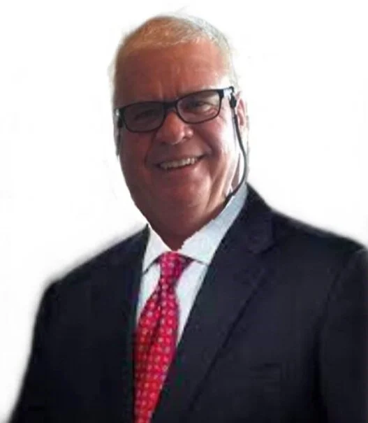 A smiling man in a dark suit, white shirt, and pink patterned tie, wearing glasses, against a plain white background.