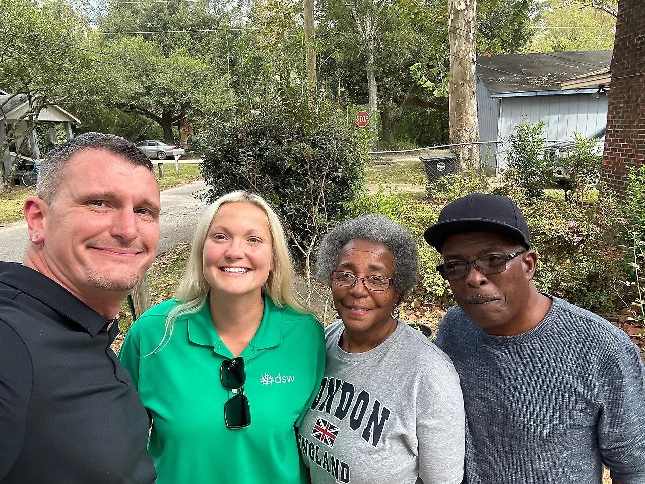 Four people standing outdoors, smiling for a photo. The background features trees, bushes, a stop sign, a building, and parked cars.