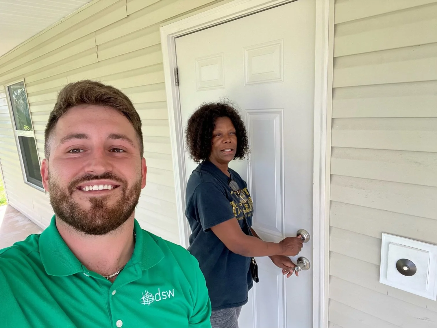 A man smiling taking a selfie in front of a woman standing at a door, holding the door handle outside a house with beige siding.