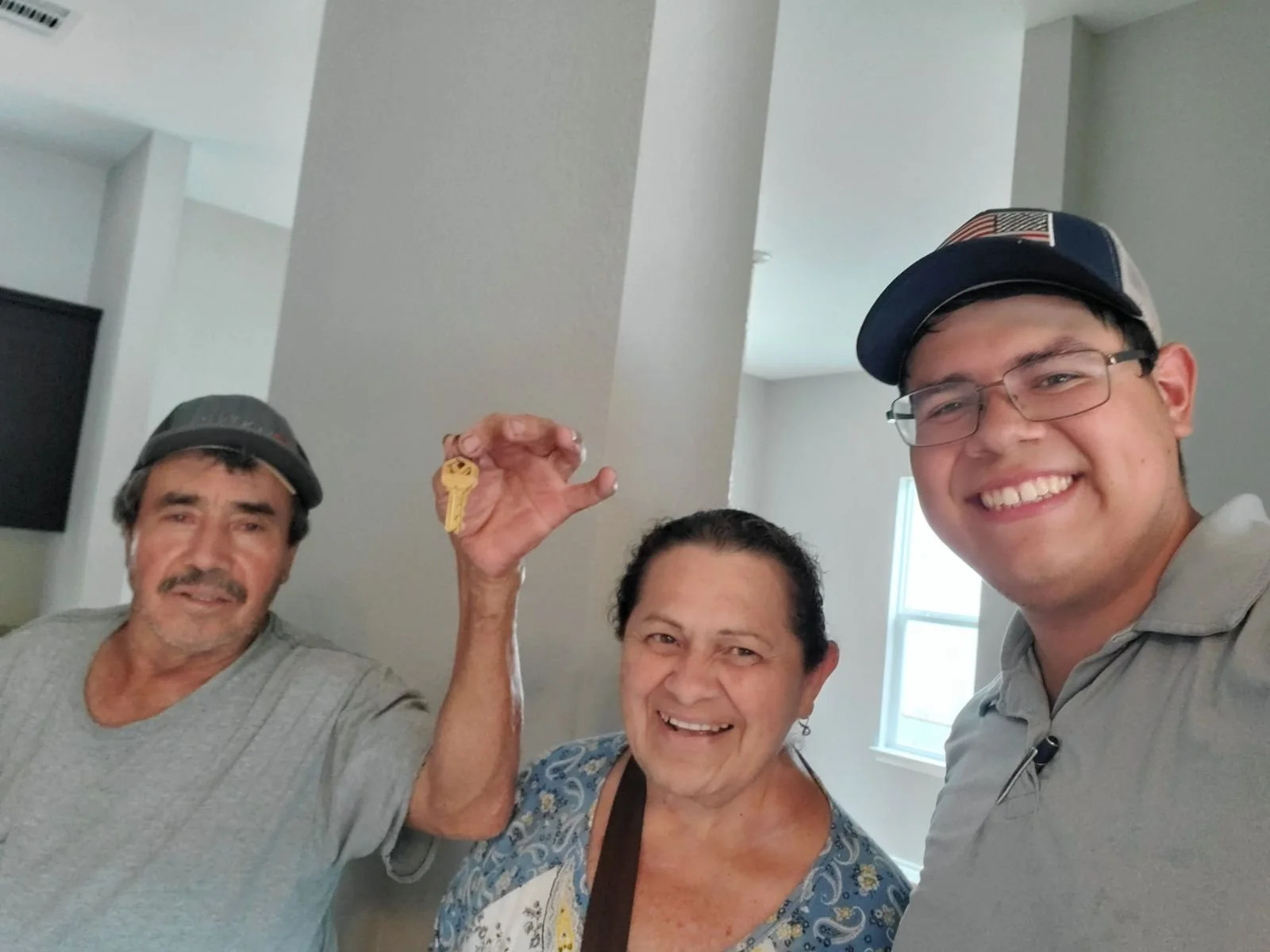 Three smiling people taking a selfie indoors, with one holding a key. An elderly man on the left, a woman in the middle, and a younger man on the right, all wearing casual clothing and caps.