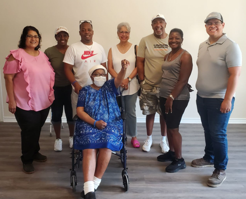 Eight people standing and one person seated in a wheelchair, all smiling indoors against a plain white wall. They are dressed casually, with one woman wearing a pink blouse and others in t-shirts and athletic wear. The seated woman is wearing a blue 