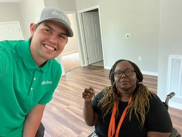 A smiling man wearing a gray cap and green polo shirt with DSW logo, standing next to a woman in glasses with braids, sitting in a wheelchair in a room with wood floors and a partially open door.