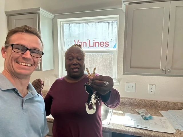 A man taking a selfie with a woman in a kitchen, holding up a set of keys. The woman has short hair and is wearing a dark shirt. The kitchen has white cabinets, granite countertops, and a window with a view of a sign that says "Van Lines" outside.