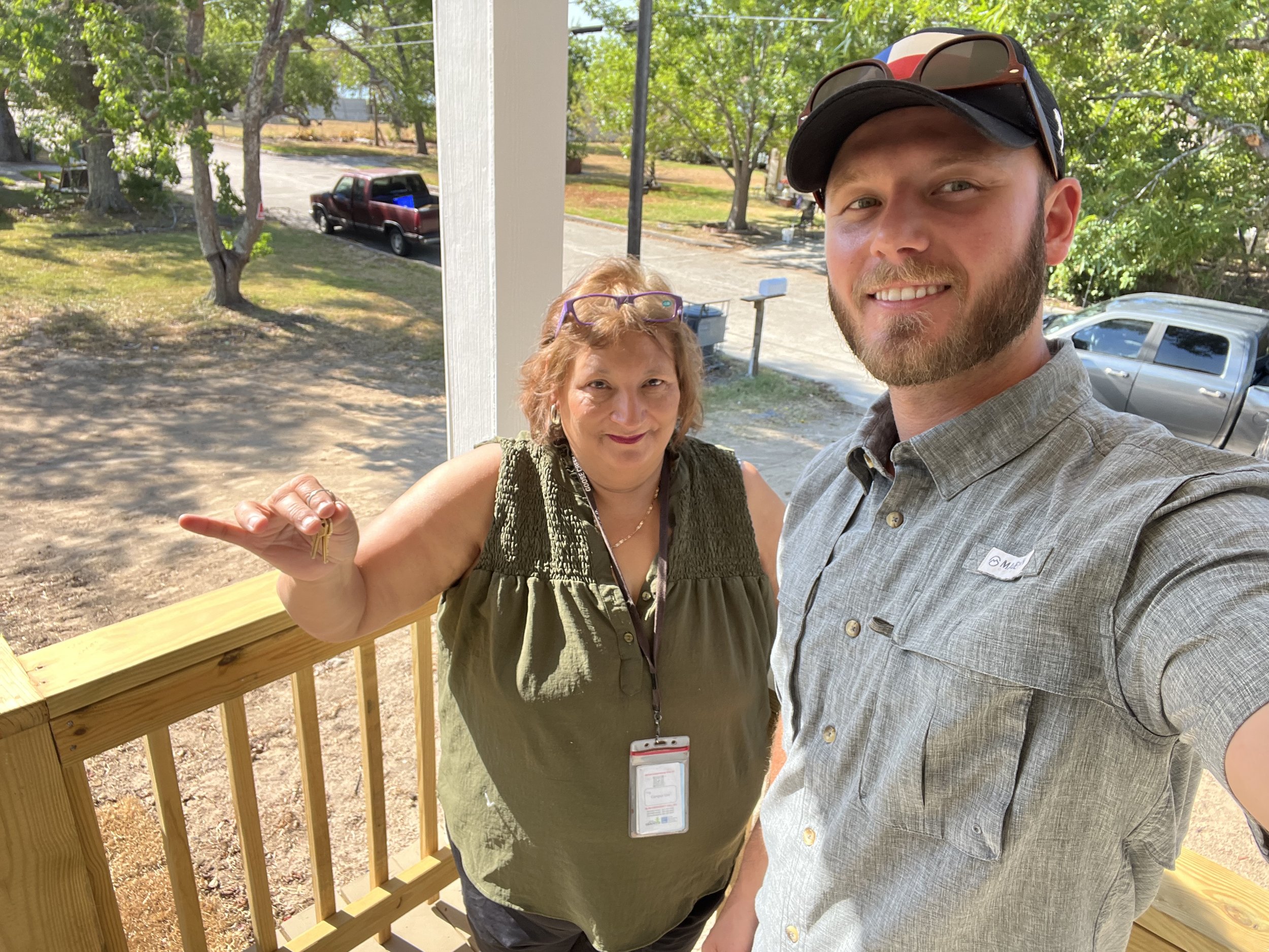A smiling young man and an older woman taking a selfie outside on a sunny day. The man is wearing a baseball cap, sunglasses, and a plaid shirt. The woman is wearing glasses, a green vest, and has a badge hanging around her neck. They are standing ne