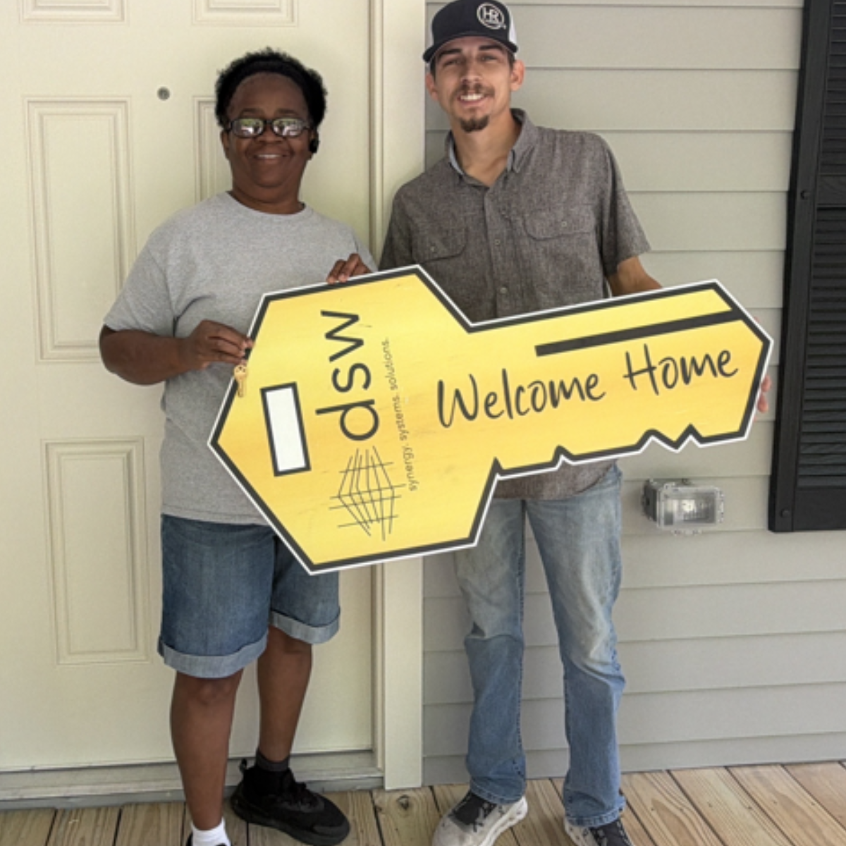 Two men standing outside near a house, holding a large yellow sign shaped like a key with the words 'dSW Welcome Home' and a logo, smiling at the camera.
