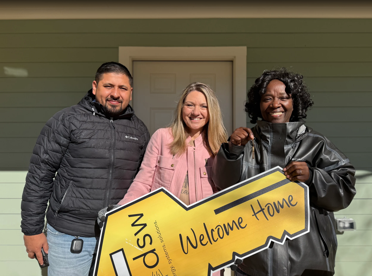 Three smiling people holding a yellow "Welcome Home" sign in front of a green house.
