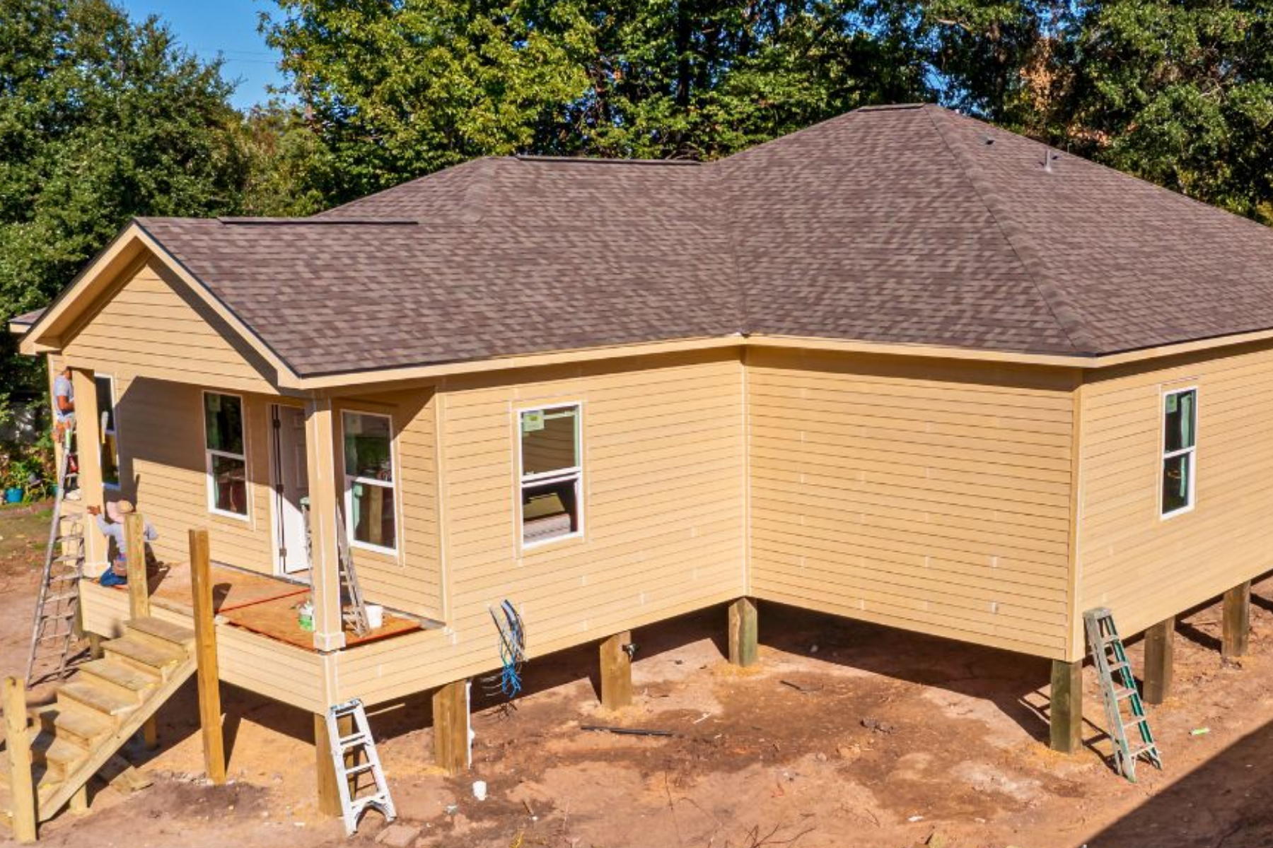 A house under construction, raised on stilts, with a yellow exterior and a brown shingle roof. Ladder and construction materials are visible around the house, with trees in the background.