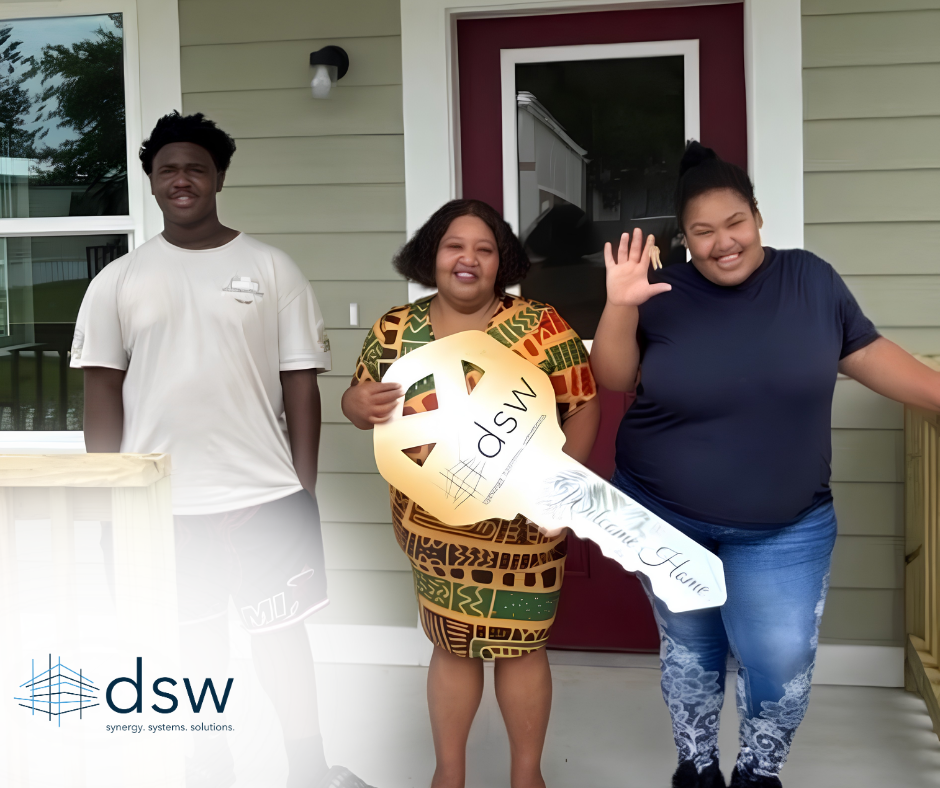 Three people standing outside a house, two women and one man. The woman in the middle is holding a large key-shaped sign with 'dsw' and 'Welcome Home' written on it. They are smiling and appear to be celebrating a new home arrival.