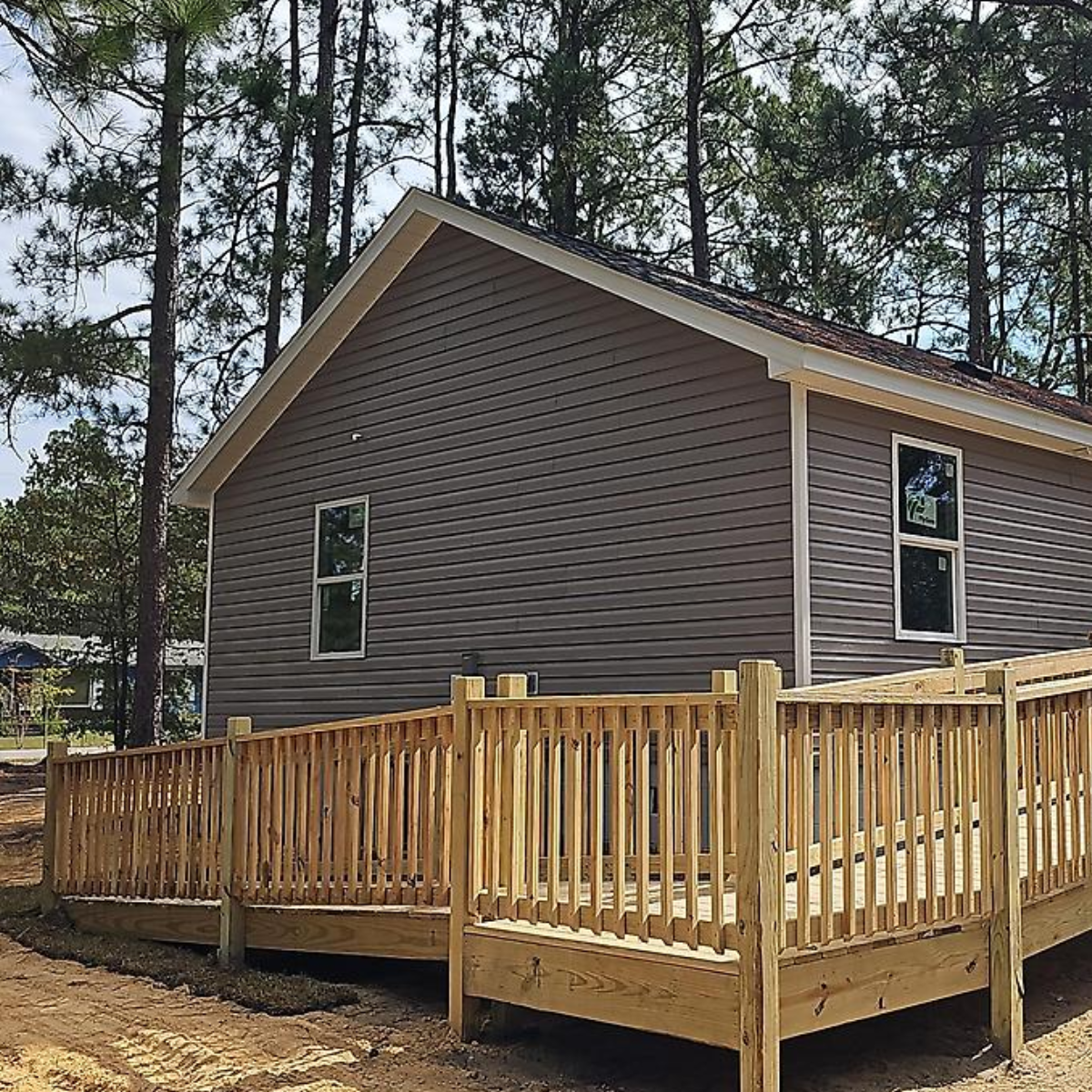 A house with gray siding and two windows, surrounded by tall pine trees. There is a new wooden deck with a railing in the foreground.