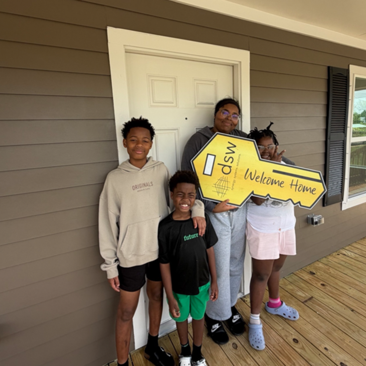 A woman and three children standing on a wooden porch in front of a house with beige siding, holding a large yellow key-shaped sign that says 'Welcome Home' and 'WSDV'.