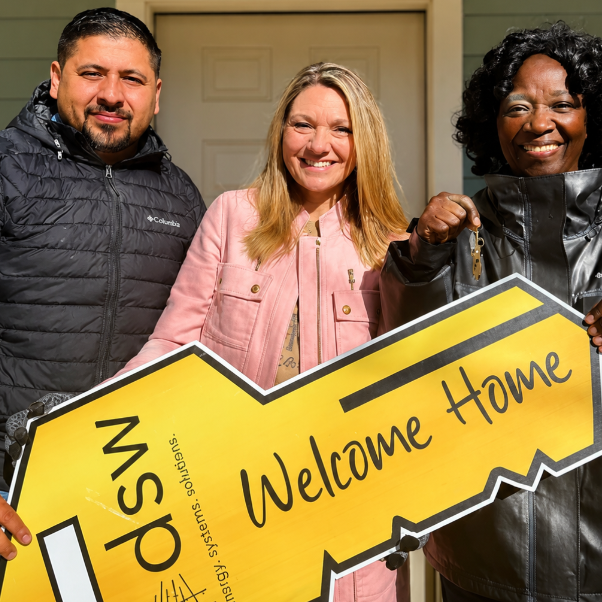 Three diverse people standing together, smiling, holding a large yellow and black 'Welcome Home' sign, outside a house.
