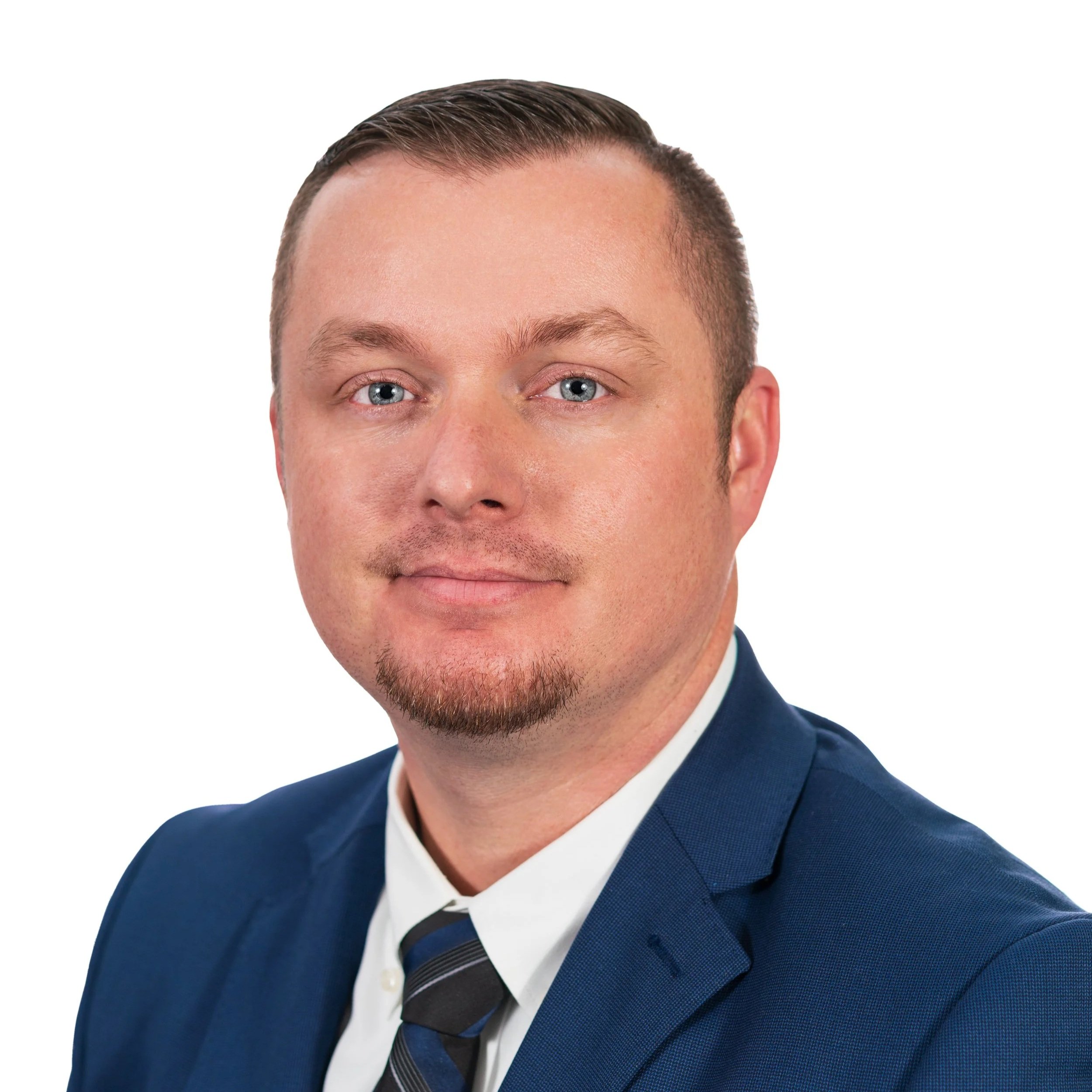 Close-up of a man wearing a navy blue suit, white dress shirt, and striped gray tie, with short brown hair and blue eyes, against a white background.