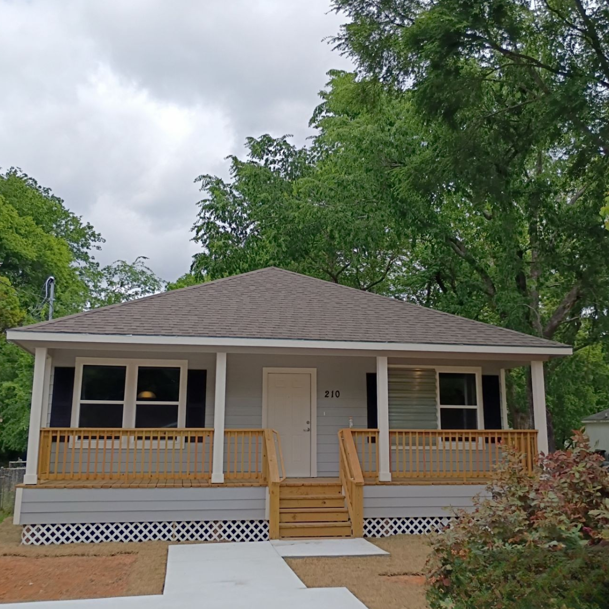 A newly built, small, white house with a front porch and stairs, located under a large leafy tree on a cloudy day.