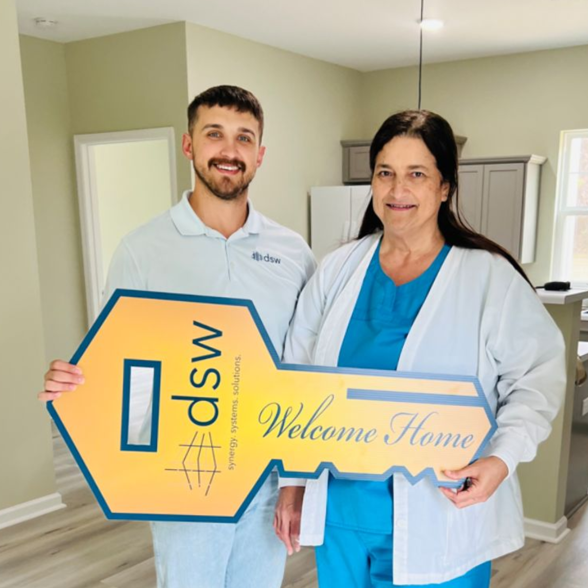 A young man and an older woman, possibly healthcare workers, holding a large yellow key-shaped sign that says 'dwsV' and 'Welcome Home' inside a home or medical facility.