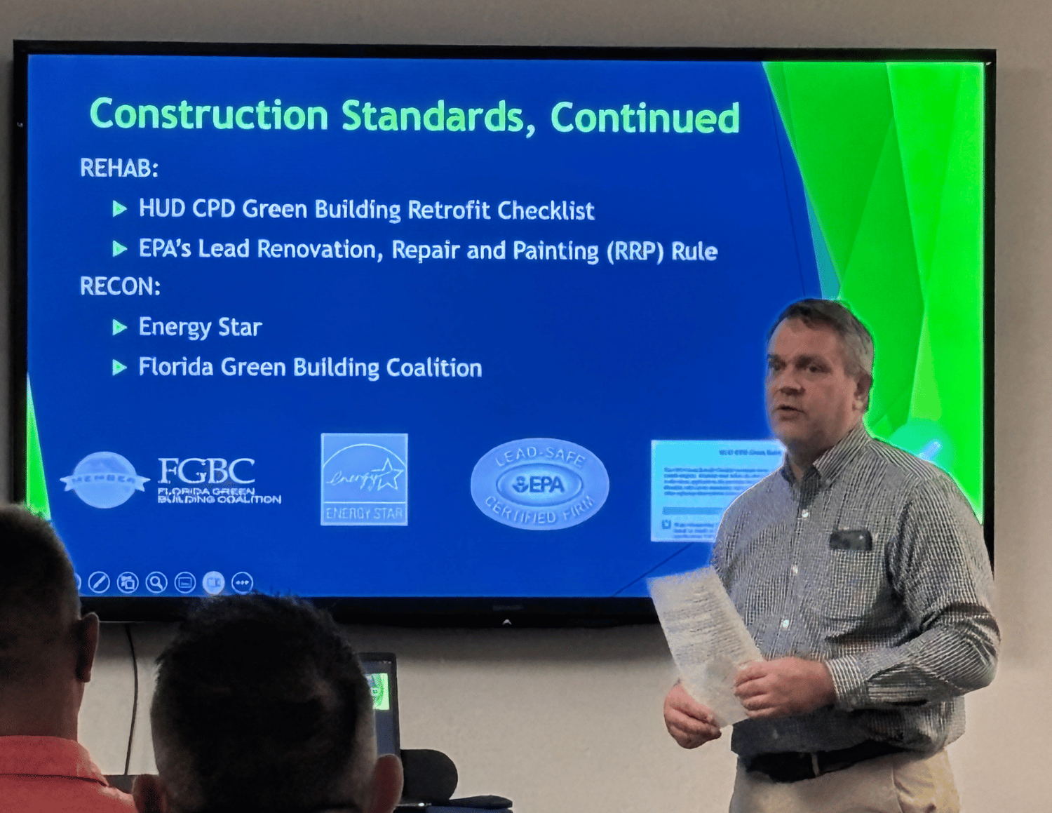 A man in a checkered shirt giving a presentation in front of a large screen displaying information about construction standards, including links to organizations like FHA, Energy Star, and EPA.