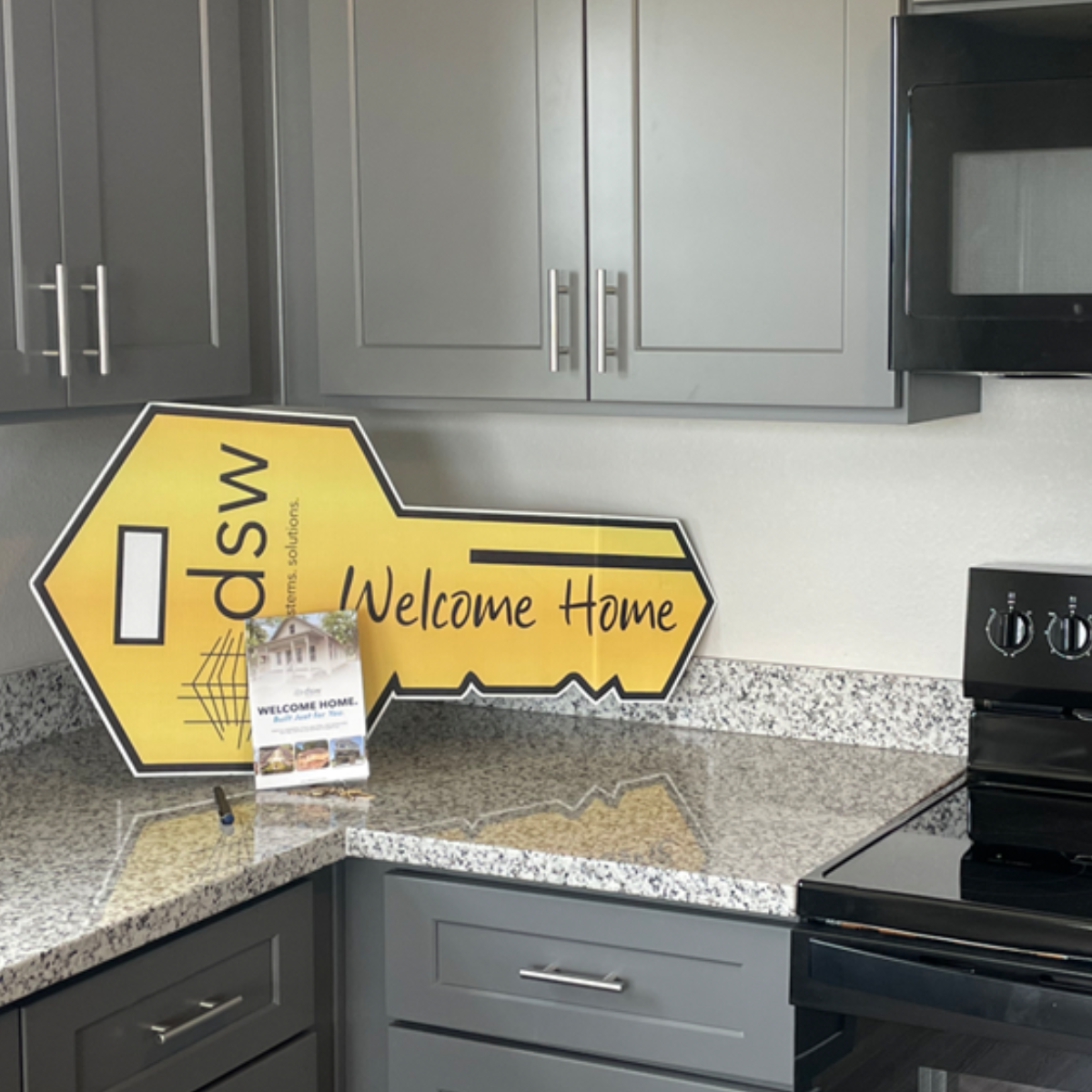 A yellow key-shaped sign with black text that reads 'SW DENTAL BEAUTY' and 'Welcome Home' is placed on a speckled granite kitchen countertop. There is also a small card or brochure with a house picture and the words 'WELCOME HOME.' The kitchen has gr