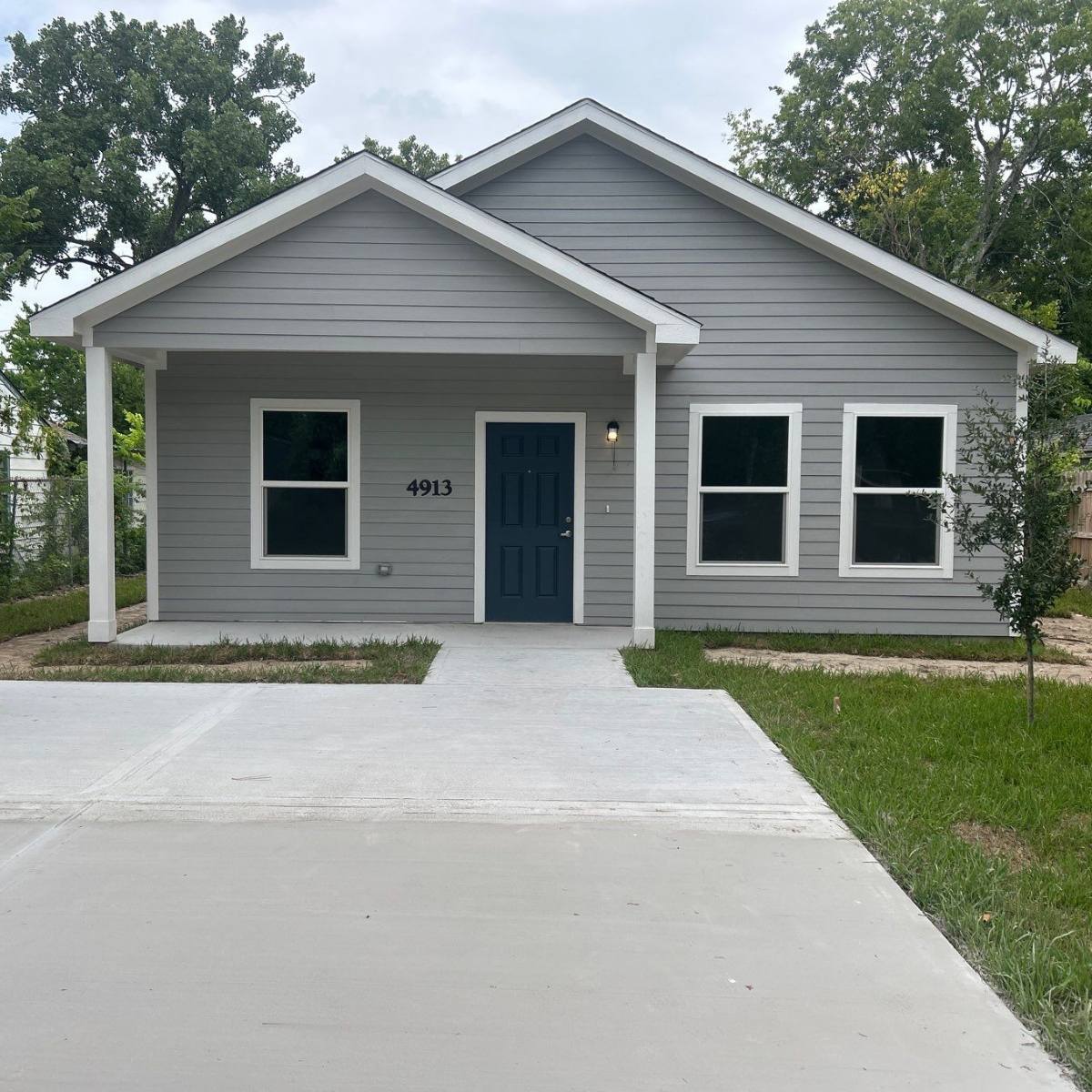 A modern gray house with white trim and a dark blue door, featuring three windows, a small front porch, and a concrete driveway, with trees in the background.