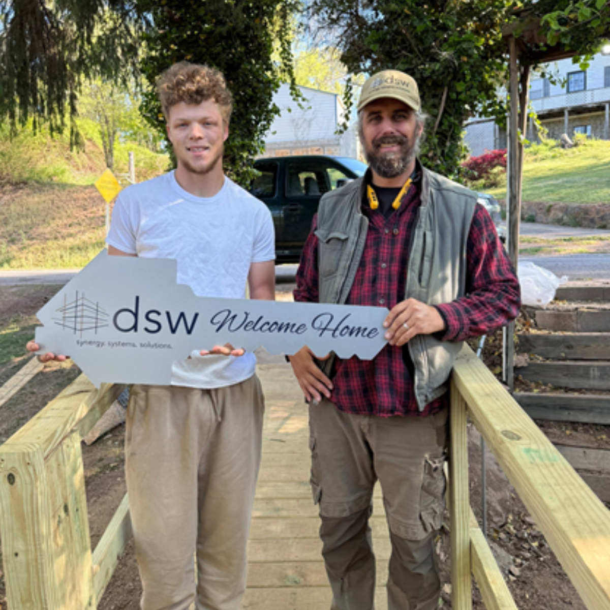 Two men standing outdoors on a wooden porch, holding a large sign that says "dsw Welcome Home" with a house outline, next to a black vehicle, trees, and a house in the background.