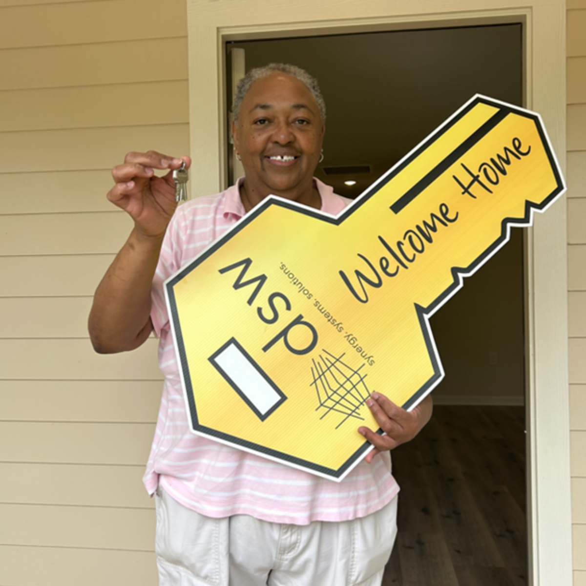 A smiling woman holding a large yellow welcome sign shaped like a key, with a key in her right hand, in front of a house door.