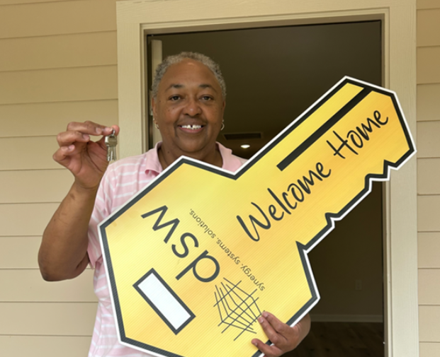Smiling woman holding a large yellow key-shaped sign that says 'Welcome Home' and 'DSM' with home automation company's name, standing inside near a doorway.