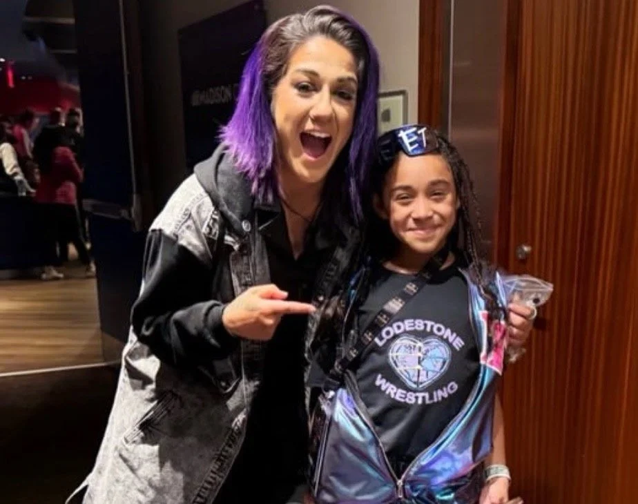 A woman with purple hair smiling and pointing at a young girl with curly hair, both standing indoors.