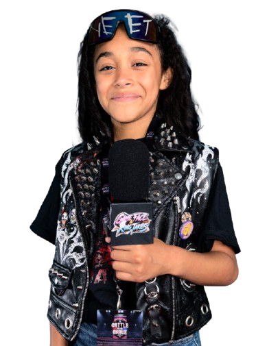 A young girl with curly hair holding a microphone with a logo, wearing sunglasses on her head and a black vest with patches, smiling.