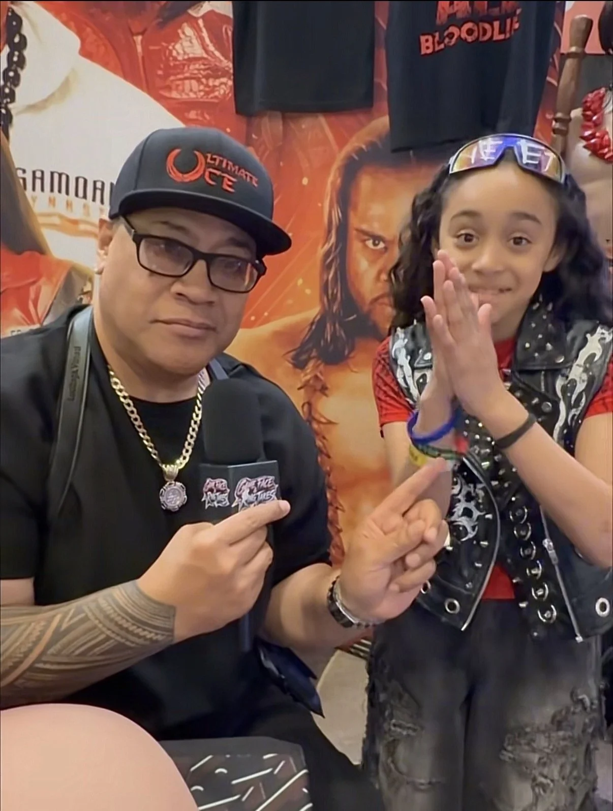 A man wearing a black cap, glasses, a black shirt, and gold chains is holding a microphone and pointing at a young girl. The girl has curly hair, sunglasses on her head, and is wearing a sleeveless vest over a red shirt. She is clasping her hands together in a prayer-like gesture, smiling at the camera, and has several colorful bracelets on her wrists. Behind them is a poster with a person's face and some clothing items hanging.