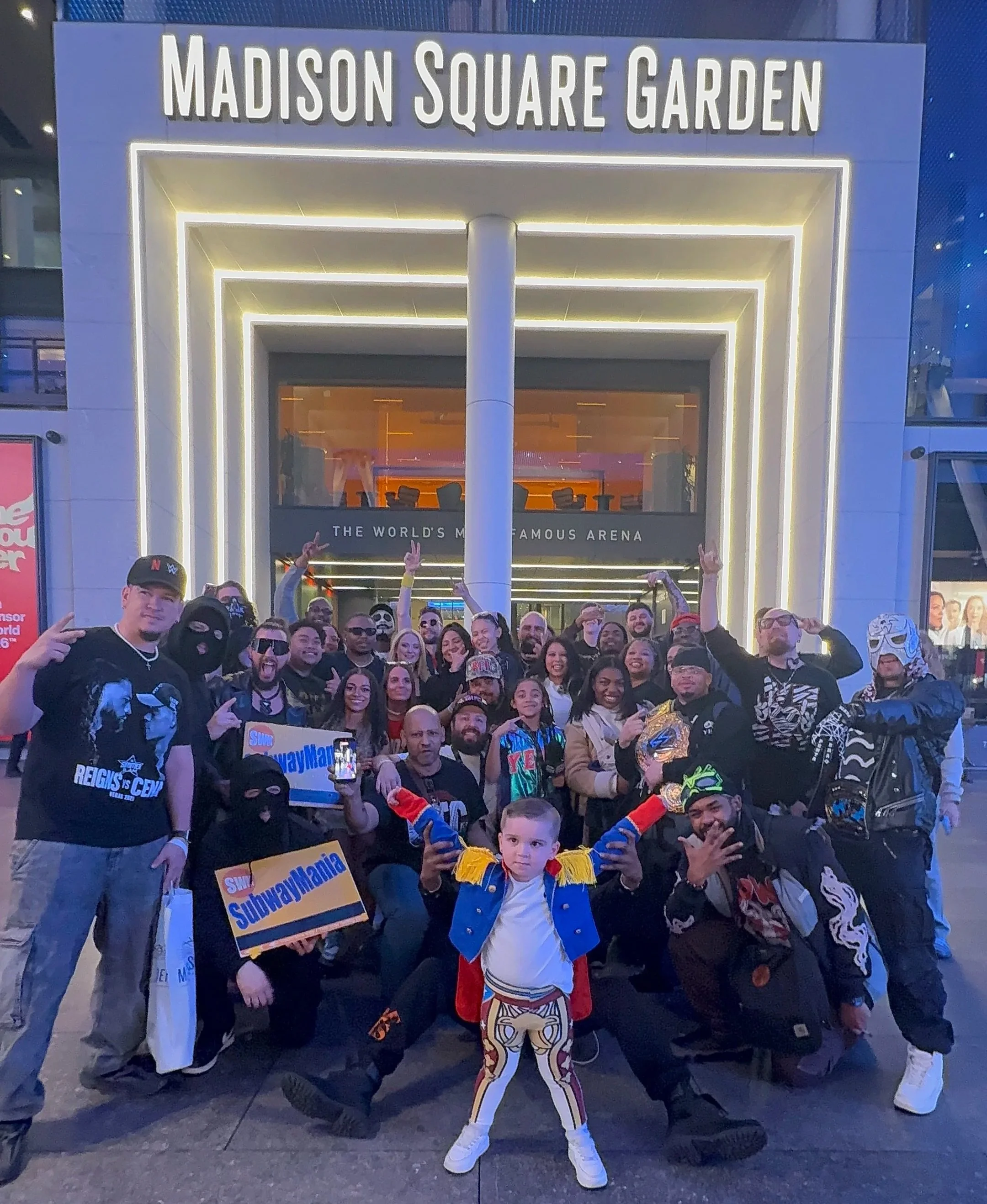 A large group of people gathered outside Madison Square Garden for Subway Mania, with a young boy in the foreground dressed in a colorful costume. Many people are holding signs and making peace signs, and the theater's entrance with illuminated signage is visible in the background.