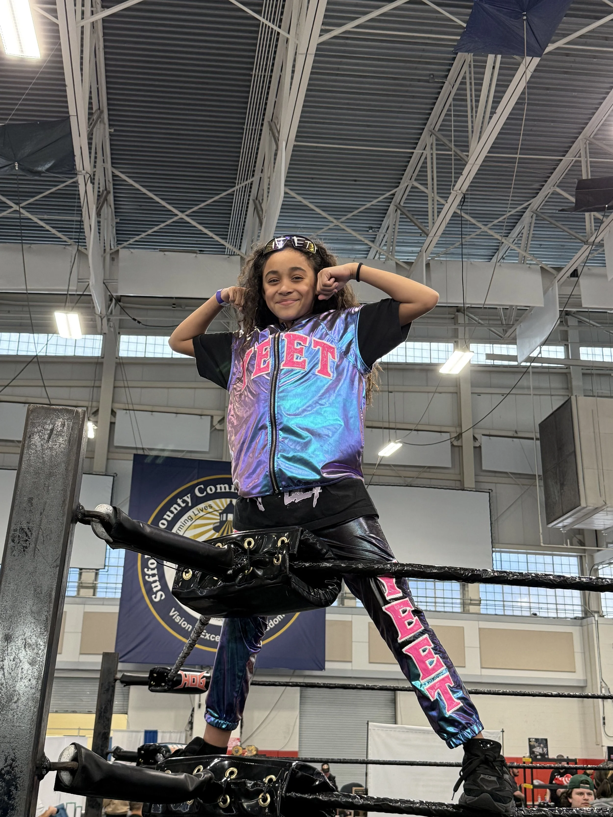 A young girl standing in a wrestling ring with her hands behind her head, wearing colorful iridescent pants and a black T-shirt that says "BEET" in pink letters. She is smiling confidently. The background shows a large indoor venue with high ceilings, industrial lighting, and judging from the banners, it appears to be a wrestling or sports event.