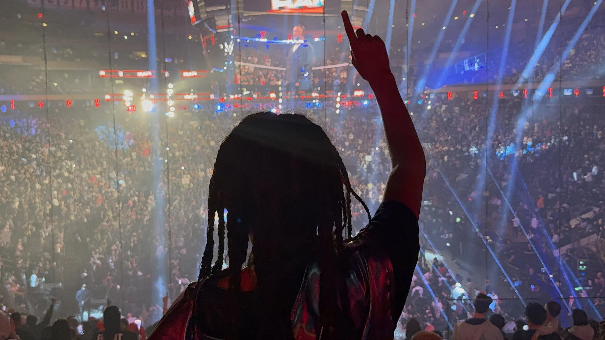 Silhouette of a person with braided hair pointing upward at a crowded concert or event with bright lights and laser effects.