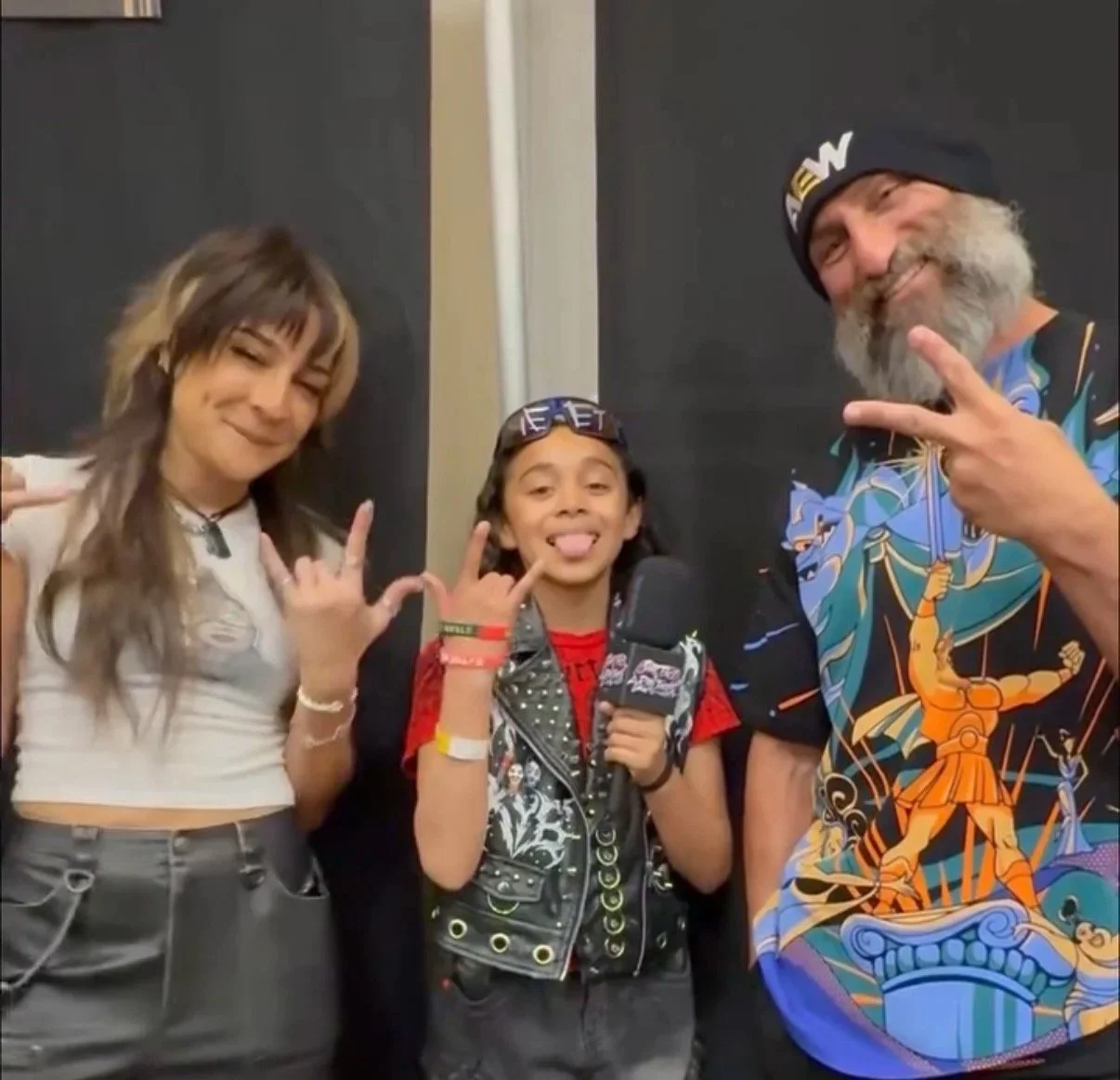Three people smiling and making rock and roll hand signs, with one person holding a microphone, indoors.