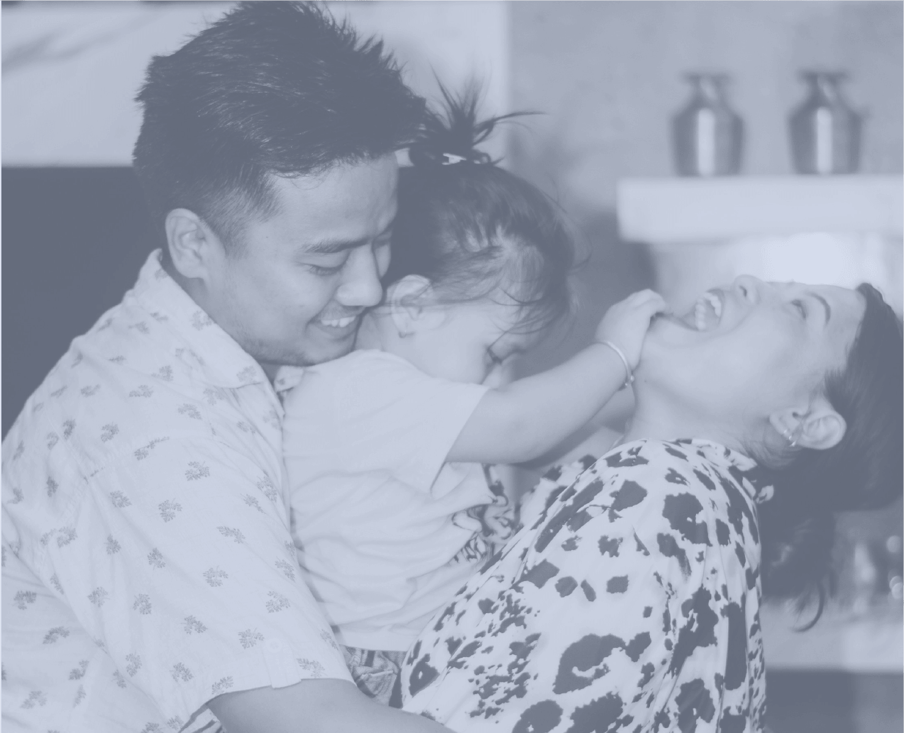 A family of three enjoying a playful moment together indoors. The father is holding a young girl, who is tickling or poking the mother, both of whom are laughing.