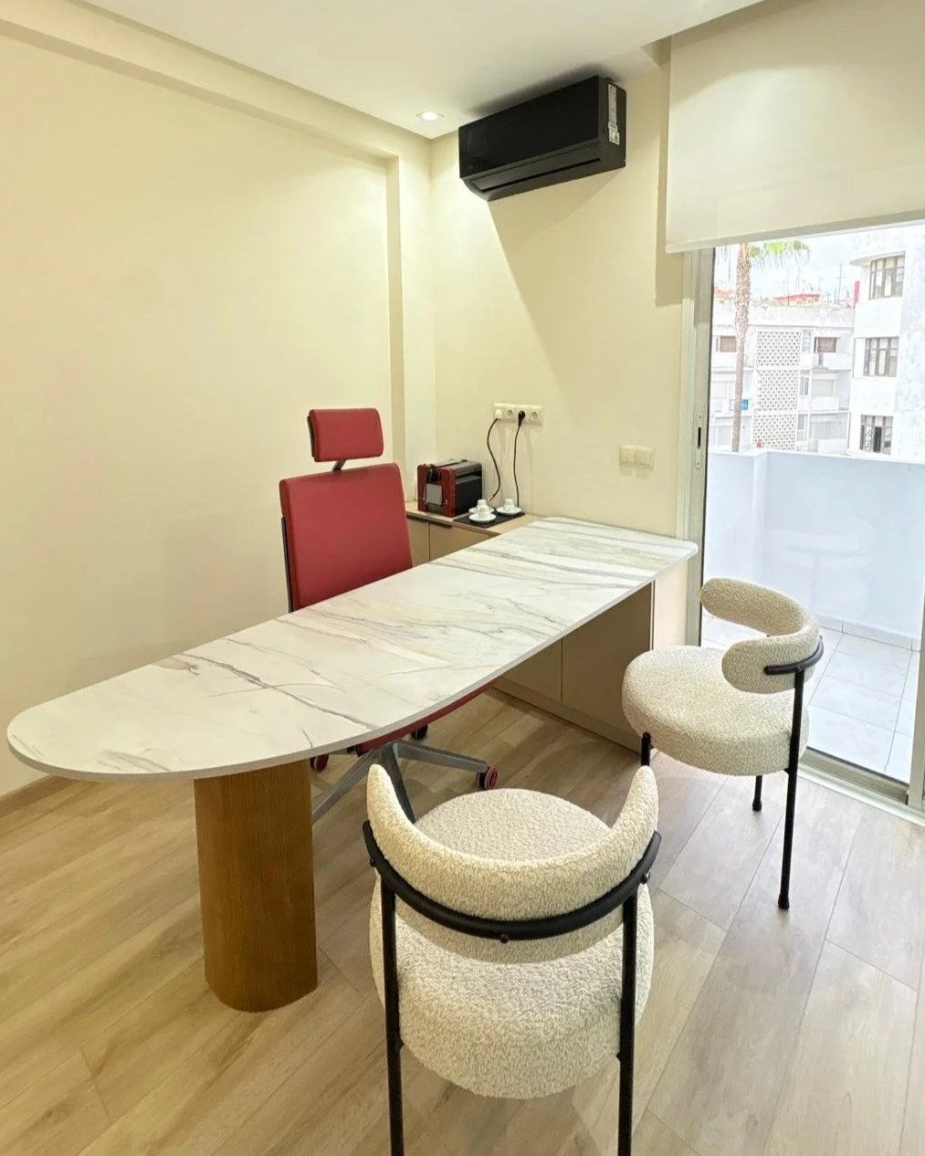 Une salle de bureau ou de réunion avec une table en marbre, une chaise rouge derrière le bureau, deux chaises beige devant, une fenêtre donnant sur un balcon, un climatiseur noir et une source d'alimentation électrique murale.