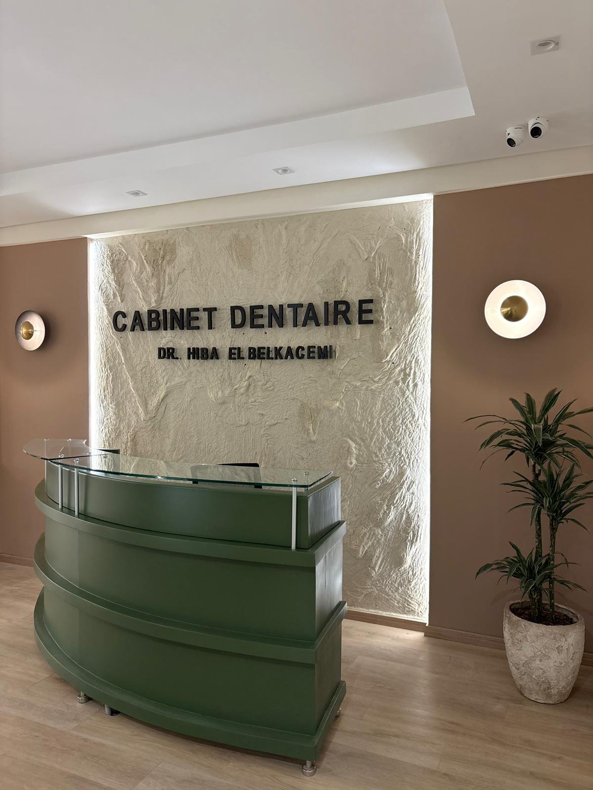Intérieur d'un cabinet dentaire avec un comptoir vert, un mur en pierre avec le nom 'CABINET DENTAIRE DR. HIBA EL BELKACEM', deux lampes murales, un plafond blanc, et une plante verte dans un pot.