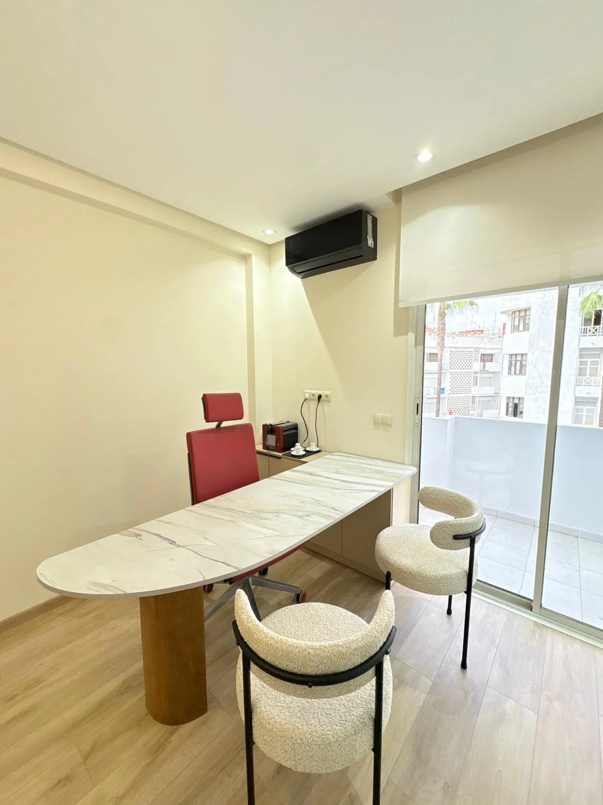 Salle de réunion avec un bureau en marbre, une chaise blanche et deux chaises de bureau rouges, une porte-fenêtre avec balcon et vue sur des immeubles, et une climatisation.
