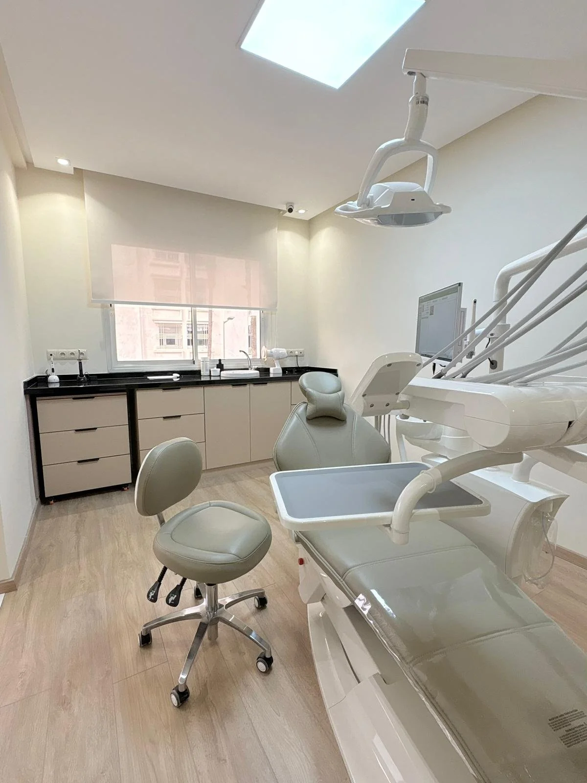 Salle de soins dentaires moderne avec un fauteuil dentaire, une lampe dentaire, un écran, un tabouret et un lavabo, dans une pièce lumineuse avec un plafond en fenêtre de toit.