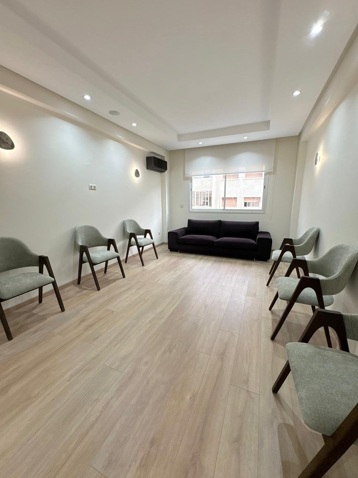 Une salle d'attente vide avec un canapé noir et plusieurs chaises en bois et tissu gris, sol en bois, murs blancs, fenêtre avec vue sur des bâtiments, éclairage encastré au plafond.