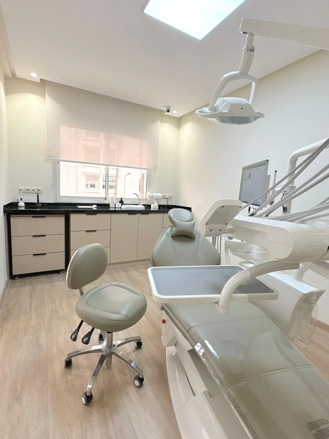 Salle de soins dentaires avec un fauteuil dentaire blanc, une chaise ergonomique beige, un ordinateur, un éclairage au plafond, un lavabo, des meubles de rangement, et une fenêtre avec vitre.
