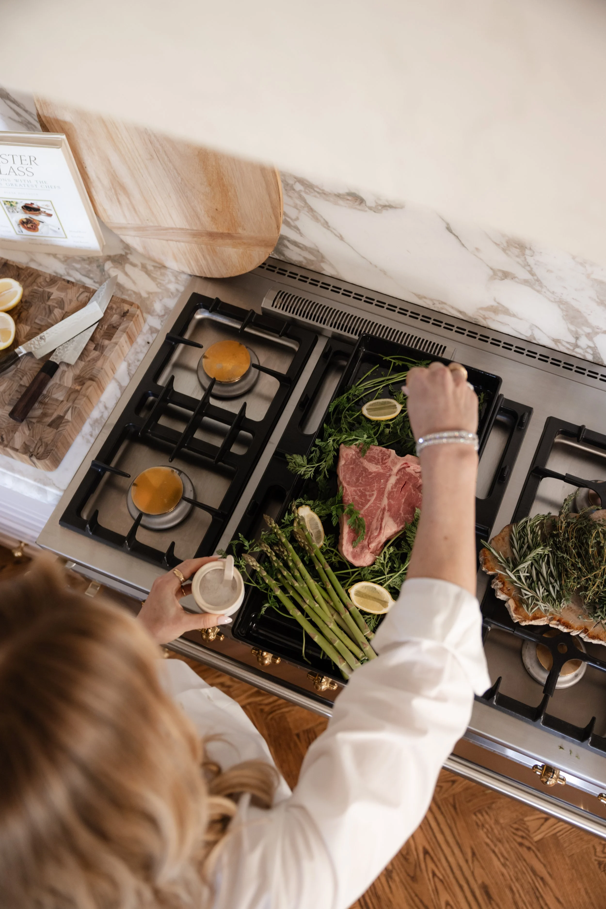 A chef cooking a steak on a cooktop beside fresh herbs and vegetables