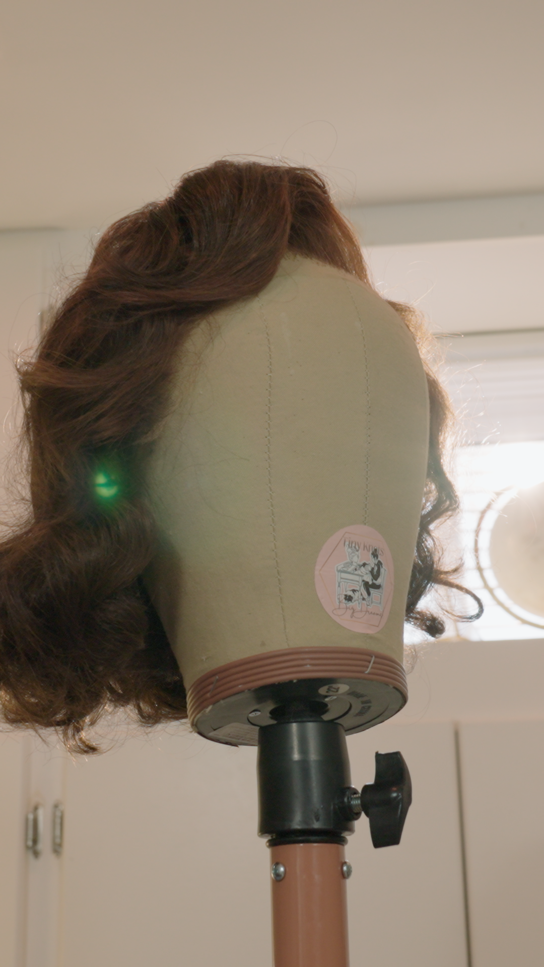 A mannequin head with long curly brown hair on a stand, with a sticker that says 'Tiny Knots Big Dreams' and an illustration of a woman at a sewing machine. Pink and Pretty is the brand with Emma Dirks, the Wig Master