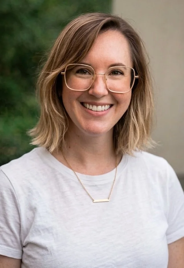 A smiling woman with shoulder-length blonde hair, wearing glasses, a white t-shirt, and a gold necklace.