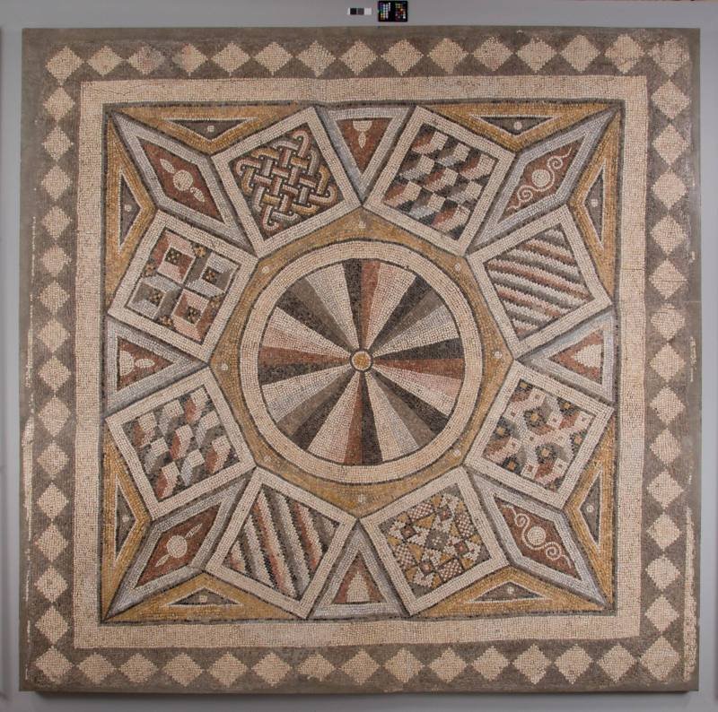 Ancient mosaic with geometric patterns, including a central circle with alternating dark and light triangular segments, surrounded by rectangular and diamond-shaped sections filled with intricate designs, all framed by a border of alternating light and dark squares.