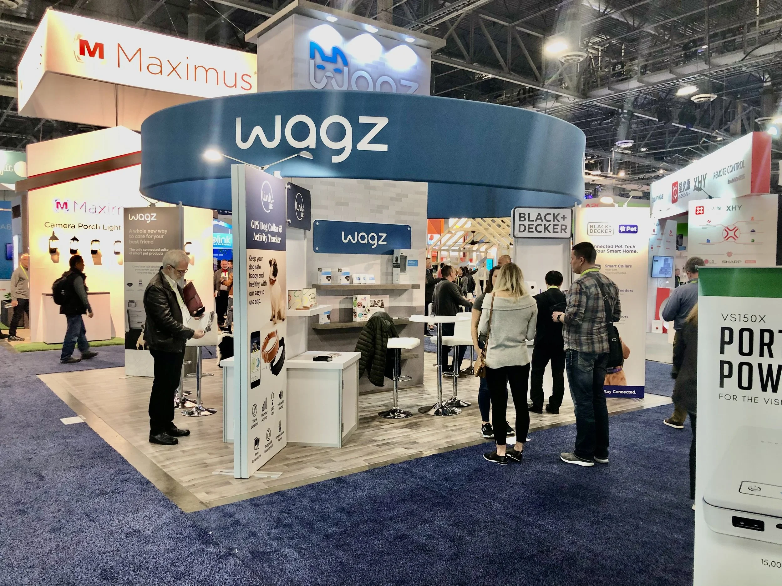 A trade show booth with the brand name 'Wagz' prominently displayed on a curved blue sign at the center. Several people are engaged, exploring the products. Surrounding booths feature brand names 'Maximus,' 'Black + Decker,' and various product displays. The venue has high ceilings with visible structural elements and lighting.