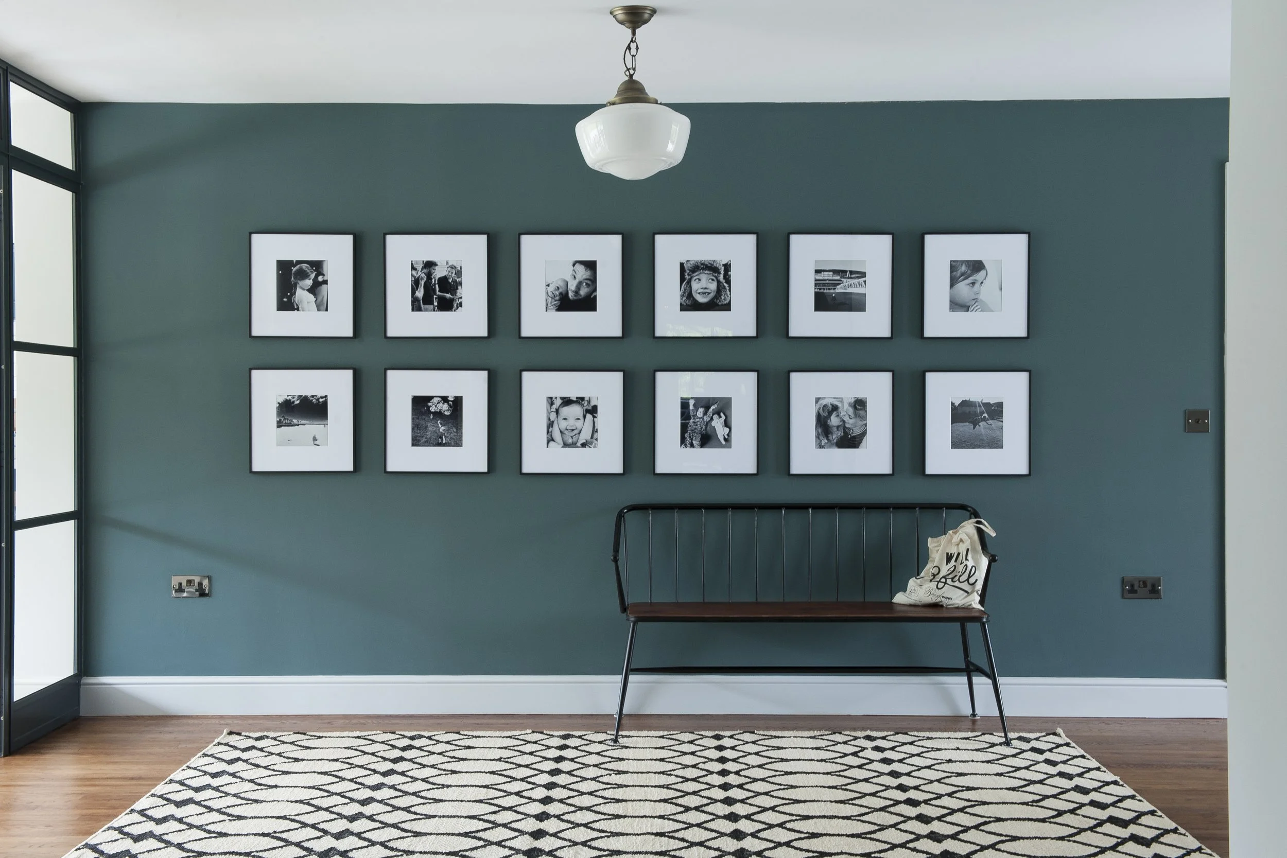 Interior of a room with teal wall decorated with black and white framed photographs of children and adults, black bench, printed tote bag, ceiling light, and a black and white patterned rug on wooden floor.