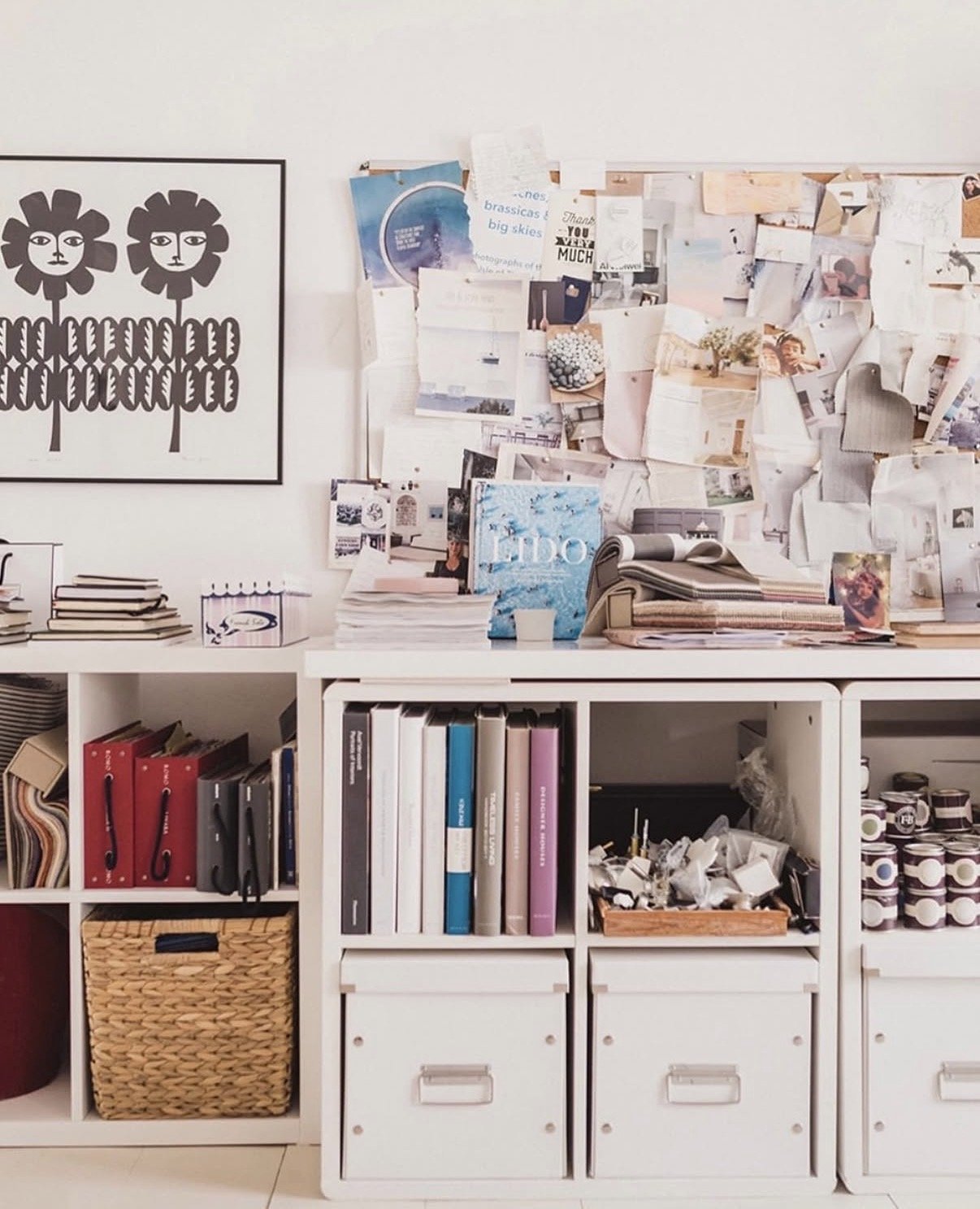 An office featuring a white desk with stacked books, papers, and folders, in front of a white shelving unit filled with binders, baskets, and supplies. Behind the desk, a large bulletin board covered with notes, photos, and papers hangs on the wall.
