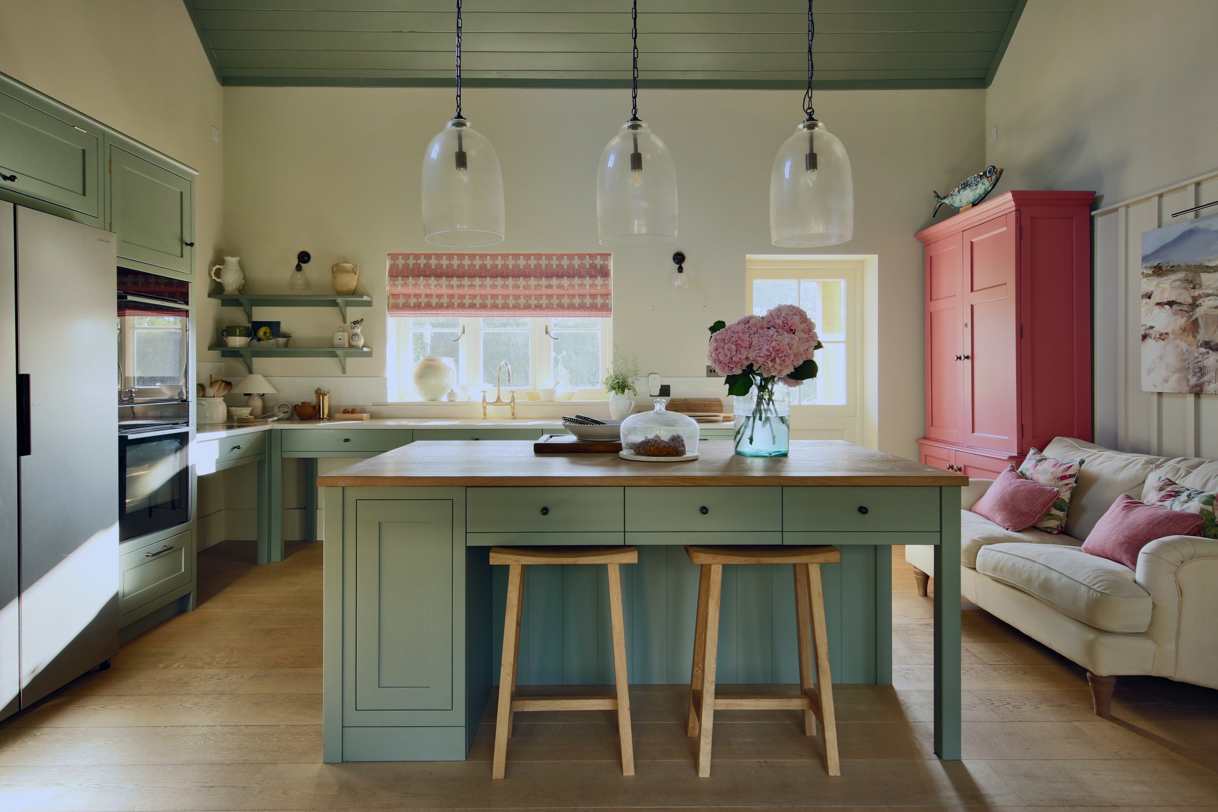 A kitchen with green lower cabinets and island, pink upper cabinet, white walls, wooden floor, large window with pink Roman shade, and a vase of pink hydrangeas on the island.