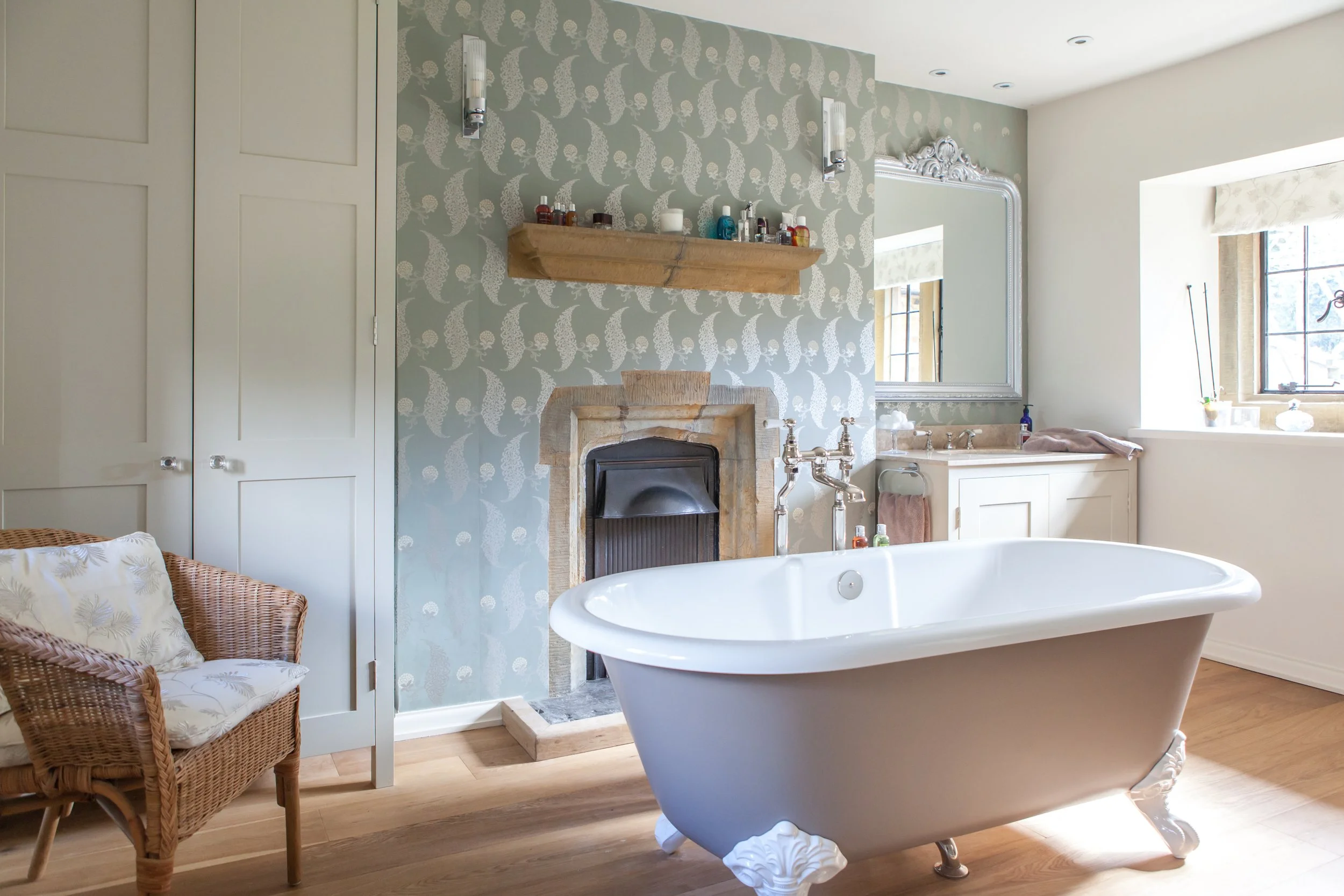 A vintage-style bathroom with a clawfoot bathtub, a stone fireplace with a wooden mantel, patterned wallpaper, a large mirror, and a window with blinds, natural light streaming in.