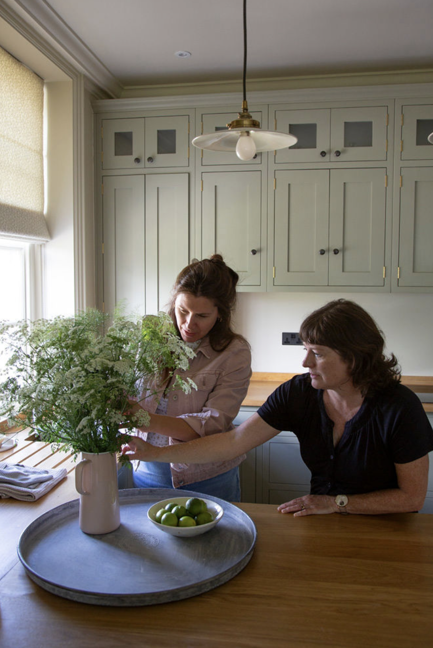 Two women arranging flowers in a kitchen with green cabinets, a large window, and a wooden countertop.